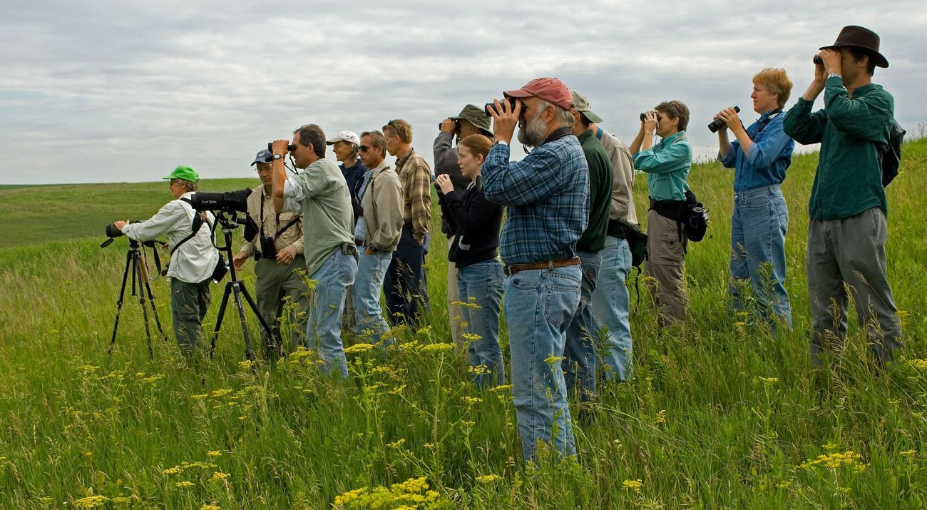 A group of people standing in a grassy field, looking into the distance, some through binoculars.