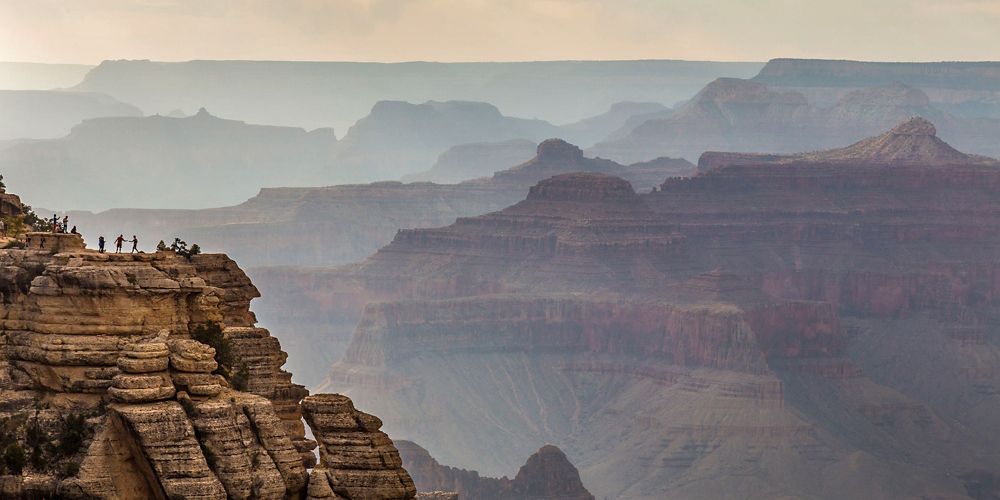 A few people stand atop a rock formation overlooking the Grand Canyon.