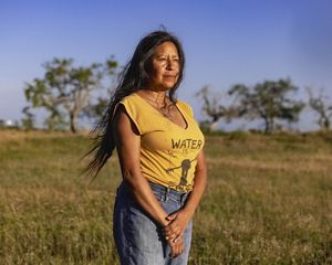 woman stands in yellow shirt, long hair, looking into the distance, brown and green grass and small trees behind her