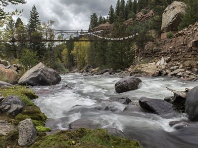 A fast-flowing river with boulders in it and along the shore. A narrow bridge crosses the gorge in the distance.