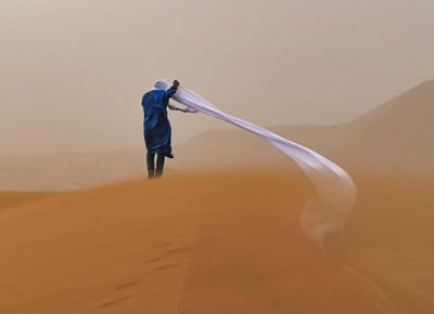 A person walks across orange sand dunes during a sandstorm and their headwrap has come undone and is blowing in the wind.